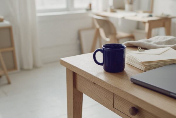 A navy mug, a stack of sealed envelopes and a closed laptop on a small business owner's oak desk