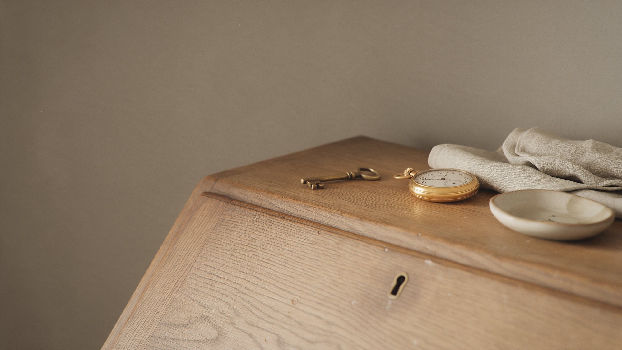 An antique brass key and a pocket watch on an old oak writing bureau beside folded linen